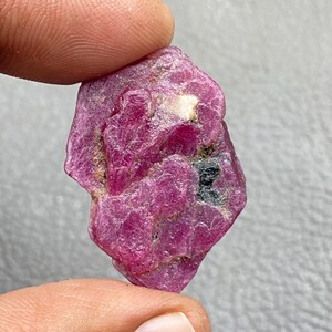 May include: A rough, uncut ruby gemstone, held between fingers. The ruby is a deep pinkish-red colour with a crystalline structure and some dark inclusions. The stone has an irregular shape and a natural, unpolished appearance.