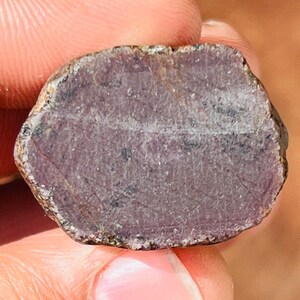 May include: A close-up of a rough, purple gemstone with a natural, irregular shape. The stone has a slightly translucent appearance and shows a hint of sparkle.