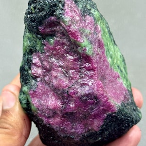 May include: A raw ruby-in-zoisite gemstone. The stone displays a vibrant magenta centre, encircled by green and black mineral deposits. The rough texture and natural form of the stone are visible.