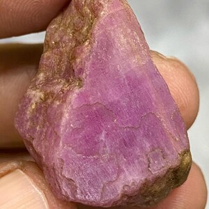 May include: A raw ruby gemstone, displaying a rich pink colour. The natural stone has a textured surface with variations of pink and some golden-brown patches.
