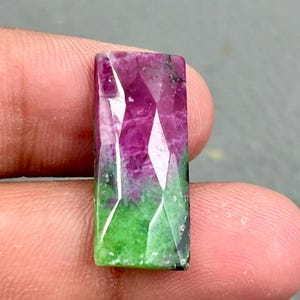 May include: A green and pink gemstone cabochon with a faceted cut. The gemstone is rectangular in shape and has a smooth, polished surface.