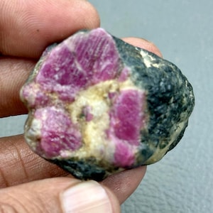 Op de afbeelding: Een ruwe, ongeslepen edelsteen met levendige magenta-kleurige robijnkristallen ingebed in een donkere, gespikkelde matrix. De robijnkristallen hebben een kristallijne structuur en de matrix heeft een mix van zwarte, grijze en bruine tinten. Deze natuursteen wordt in een hand gehouden.