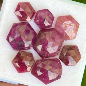 May include: A collection of eight faceted ruby gemstones, each with a unique shape and size. The rubies are a deep red color with a slight pink hue. They are arranged in a random pattern on a white surface.