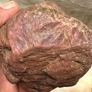 Könnte beinhalten: Ein rotbrauner Felsen mit strukturierter Oberfläche. Der Felsen weist eine Mischung aus dunklen und hellen Rot- und Brauntönen mit sichtbaren Graten und Spalten auf. Die Gesamtform ist unregelmäßig und natürlich, was auf ein rohes, unpoliertes Mineralexemplar hindeutet.