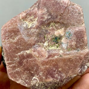 May include: A large, rough, pink ruby crystal with a natural, unpolished surface. The crystal has a deep pink colour with some white and green inclusions.