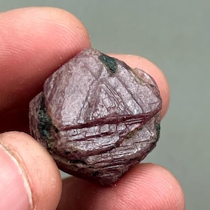 May include: A deep red ruby crystal with a faceted, geometric shape. The crystal has a rough, natural texture and is a vibrant shade of red.