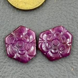 Könnte beinhalten: Zwei sechseckige, geschnitzte Edelsteine mit floralem Design. Die Edelsteine haben eine tiefmagentafarbene Farbe und eine polierte Oberfläche. Das florale Design ist zentriert mit nach außen strahlenden Blütenblättern. Im Hintergrund ist eine Goldmünze zu sehen.