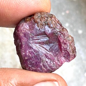 Può includere: Una pietra preziosa di rubino rosa scuro grezza con una finitura naturale non lucidata. Il rubino ha un colore profondo e ricco e un aspetto leggermente traslucido.