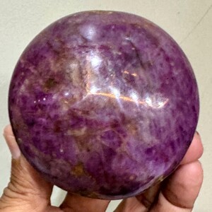 May include: A polished, spherical stone with a deep purple and magenta hue, featuring natural mottled patterns, internal crystalline textures, and light-reflecting surfaces throughout the rounded body of the mineral specimen.