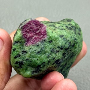 Op de afbeelding: Een ruwe edelsteen met een levendige robijninsluiting in een groene zoisietmatrix. De zoisiet is bespikkeld met zwarte toermalijn, wat een opvallend contrast creëert. De robijn heeft een diepe, rijke rode kleur.
