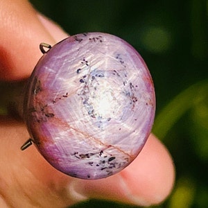 May include: A round, polished gemstone with a star-like effect. The gem is primarily a deep pink color with white and dark gray inclusions. A small metal loop is attached to the top of the stone.
