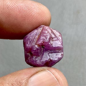 Peut inclure: Une pierre précieuse facettée, de couleur magenta foncé, tenue entre deux doigts. La gemme a une forme géométrique avec des lignes et des facettes sculptées. La surface de la pierre reflète la lumière, soulignant sa structure cristalline.