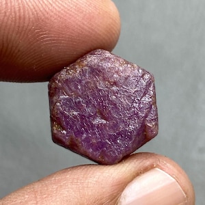 May include: A raw, hexagonal ruby gemstone. The gem is a deep, saturated purplish-red colour with a slightly textured surface. The ruby is held between two fingers against a neutral background.
