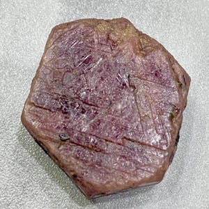 May include: A natural, rough-cut ruby gemstone. The ruby displays a deep pinkish-red colour with visible surface textures and natural inclusions. The stone's irregular shape and facets catch the light, showcasing its raw beauty. A gemstone for jewellery making.