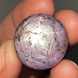May include: A close-up of a purple star sapphire cabochon with a six-pointed star pattern. The stone has a silvery sheen and is set in a silver bezel.
