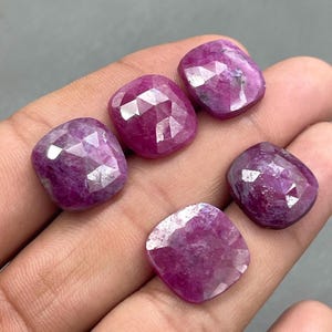 Puede incluir: Cinco rubíes facetados, tallados en cojín, en tonos rosados y rojos. Las piedras preciosas se sostienen en una mano, mostrando sus superficies pulidas y facetas geométricas. Los rubíes son una exhibición vibrante de belleza natural.