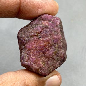 Könnte beinhalten: Ein rauer, ungeschliffener Rubin-Edelstein mit tiefroter Farbe. Der Naturstein hat eine strukturierte Oberfläche und eine unregelmäßige Form, die seinen rohen, unpolierten Zustand zeigt. Der Rubin wird zwischen zwei Fingern gehalten.
