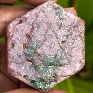 Puede incluir: Una gema hexagonal en bruto, de color rosa-púrpura. La piedra tiene áreas de inclusiones verdes y marrones, y una textura superficial visible. La gema se sostiene en una mano, mostrando su apariencia natural y sin pulir. El tema general son las gemas en bruto.