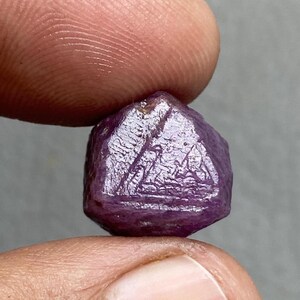 May include: A faceted, deep purple gemstone with a rough, textured surface. The gem is held between two fingers, showcasing its natural, unpolished state. The stone's color ranges from dark violet to a reddish-purple hue.