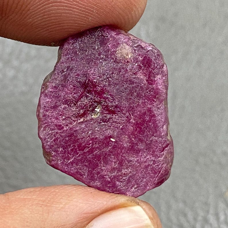 May include: A rough, uncut ruby gemstone with a deep magenta color. The surface has a textured, uneven appearance, with visible crystalline structures and natural imperfections. The gemstone is held between fingers.