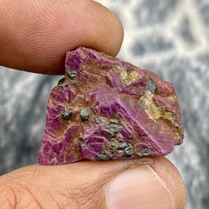 May include: A rough-cut ruby gemstone, held between fingers. The ruby displays a deep red-purple hue with visible crystalline structures and flecks of black and gold. The raw mineral has a natural, unpolished appearance.