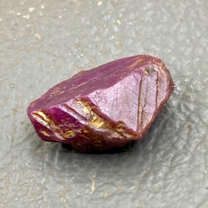 May include: A rough-cut ruby gemstone with a deep red hue and golden inclusions. The natural stone has a faceted appearance with visible lines and a slightly irregular shape. The ruby is displayed against a neutral background.