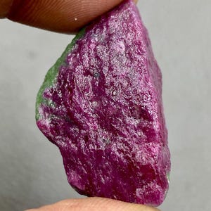 Puede incluir: Una gema de rubí sin cortar en bruto con un tono rosa intenso e inclusiones verdes. La gema se sostiene en una mano, mostrando su forma y textura naturales.