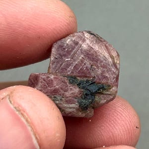 Può includere: Primo piano di un cristallo di rubino rosa con inclusioni nere. Il cristallo è sfaccettato e ha una texture ruvida e naturale.