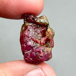 May include: A rough, red ruby gemstone with a dark red color and a slightly metallic sheen. The ruby has a natural, unpolished appearance.