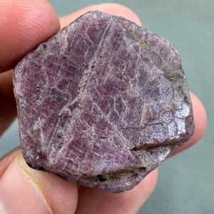 Puede incluir: Una gema de rubí en bruto, sin cortar, con un tono rojo púrpura intenso. La superficie muestra texturas naturales y estructuras cristalinas, con líneas e imperfecciones visibles. La gema se sostiene en una mano.