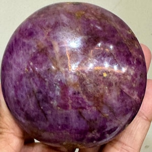 May include: A polished, spherical stone with a deep purple and magenta colour palette. The surface features natural variations, including dark brown veining, translucent patches, and small, reflective golden-toned inclusions throughout the orb.