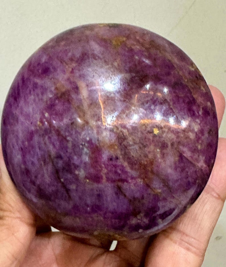 May include: A polished, spherical stone with a deep purple and magenta colour palette. The surface features natural variations, including dark brown veining, translucent patches, and small, reflective golden-toned inclusions throughout the orb.
