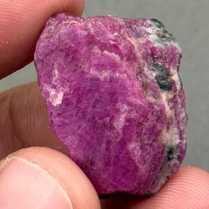 Könnte beinhalten: Ein roher, ungeschliffener Rubin-Edelstein. Der Rubin hat eine tiefe magentafarbene Farbe mit Bereichen in hellerem Rosa und Dunkelgrau. Die Oberfläche hat ein strukturiertes, kristallines Aussehen. Der Edelstein wird zwischen zwei Fingern gehalten.