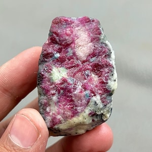 May include: A rough-cut gemstone with a vibrant, deep red hue, interspersed with lighter pink and white patches. The stone has a textured surface with hints of green and blue, suggesting a natural mineral formation. The overall shape is irregular, showcasing its raw, unpolished state.