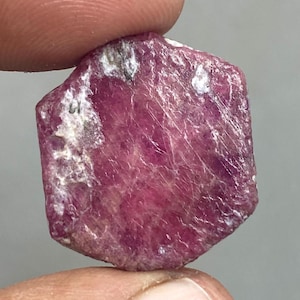 May include: A rough ruby gemstone, displaying a deep pinkish-red colour. The surface has a textured appearance with white and grey inclusions. The gemstone is held between two fingers, showing its natural, irregular shape.