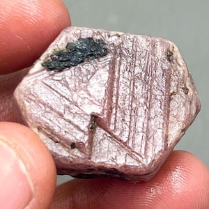 May include: A pink ruby crystal with a hexagonal shape and a dark grey inclusion. The crystal has a smooth, polished surface and is a beautiful example of a natural gemstone.