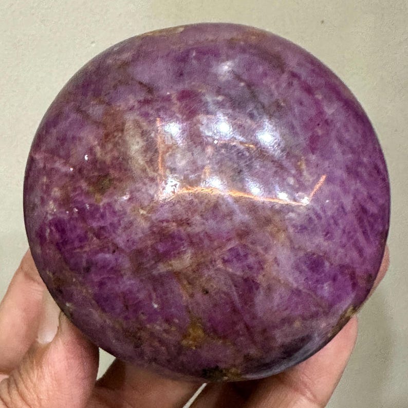 May include: A polished, spherical gemstone with a deep magenta and purple hue, featuring natural internal veining, crystalline textures, and varied opaque and translucent patches throughout its smooth, rounded surface.