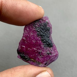 Könnte beinhalten: Ein roher Rubin-Edelstein mit tiefmagentafarbener Tönung und schwarzen Einschlüssen. Das rohe Mineral wird zwischen Fingern gehalten und zeigt seine natürliche, unpolierte Textur. Der Rubin ist ein lebendiges Beispiel für einen Naturstein.