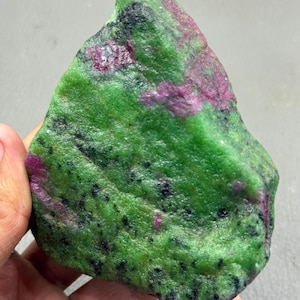 May include: A rough-cut gemstone with a vibrant green base color, speckled with black and accented by patches of deep red. The stone is held in a hand, showcasing its natural, unpolished texture. The overall appearance suggests a raw mineral specimen.