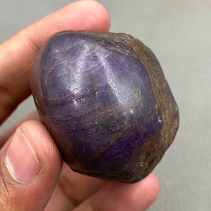 以下が含まれることがあります： 手で持たれた、深く紫色の研磨された天然宝石。 石は滑らかで丸みを帯びた形状で、表面の質感と色の変化が見え、濃い紫色から茶色まであります。