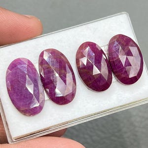 Puede incluir: Tres piedras preciosas ovaladas facetadas en una caja de exhibición blanca. Las piedras preciosas son de color rosa intenso con un ligero tono púrpura.