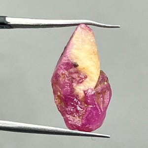 Könnte beinhalten: Ein roher Rubin-Edelstein in Pink und Weiß mit einer spitzen Spitze. Der Rubin wird von einer Pinzette gehalten.