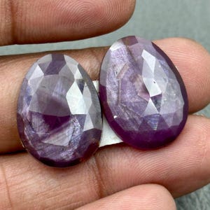 May include: Two oval-shaped, faceted gemstones in a deep purple colour. The gems have a polished, reflective surface with internal patterns. The stones are held between fingers, showing their size and colour.