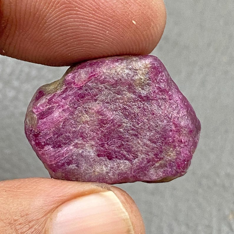 May include: A rough, uncut ruby gemstone. The ruby is a deep, vibrant pinkish-red color with a slightly uneven surface. The stone is held between two fingers, showcasing its natural, raw form.