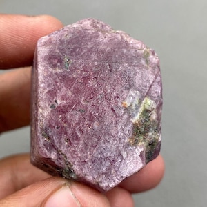May include: A raw ruby crystal, displaying a rich pink colour. The surface is textured, with patches of green and darker inclusions. The gemstone is held in a hand, highlighting its natural, unpolished state.
