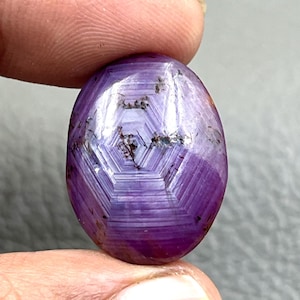 Könnte beinhalten: Ein ovaler Cabochon-Edelstein in tiefem Violett mit einem deutlichen, hellen, sechseckigen Sternmuster in der Mitte. Der Stein weist sichtbare natürliche Einschlüsse auf und besitzt eine glatte, polierte Oberfläche.