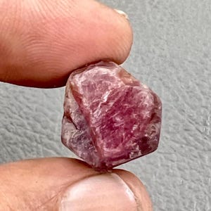 May include: A raw, uncut ruby gemstone held between fingers. The ruby has a deep pinkish-red hue with visible crystalline structures and a slightly rough texture. The gemstone is a natural, unpolished specimen.