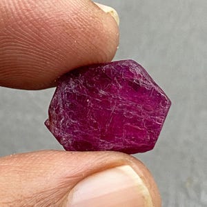 May include: A deep red, raw ruby gemstone, held between two fingers. The ruby has a rough, textured surface and a hexagonal shape. The gemstone is a rich, saturated colour and appears unpolished.