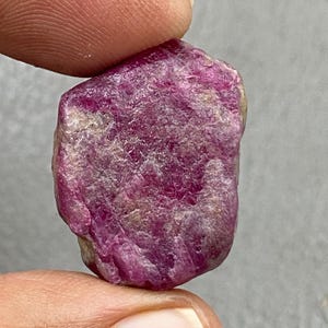 May include: A rough, uncut ruby gemstone with a deep pinkish-red hue. The surface has a textured, irregular appearance, with areas of lighter color. The gemstone is held between two fingers, showcasing its natural, raw form.