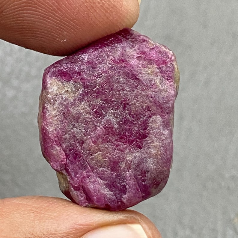 May include: A rough, uncut ruby gemstone with a deep pinkish-red hue. The surface has a textured, irregular appearance, with areas of lighter color. The gemstone is held between two fingers, showcasing its natural, raw form.